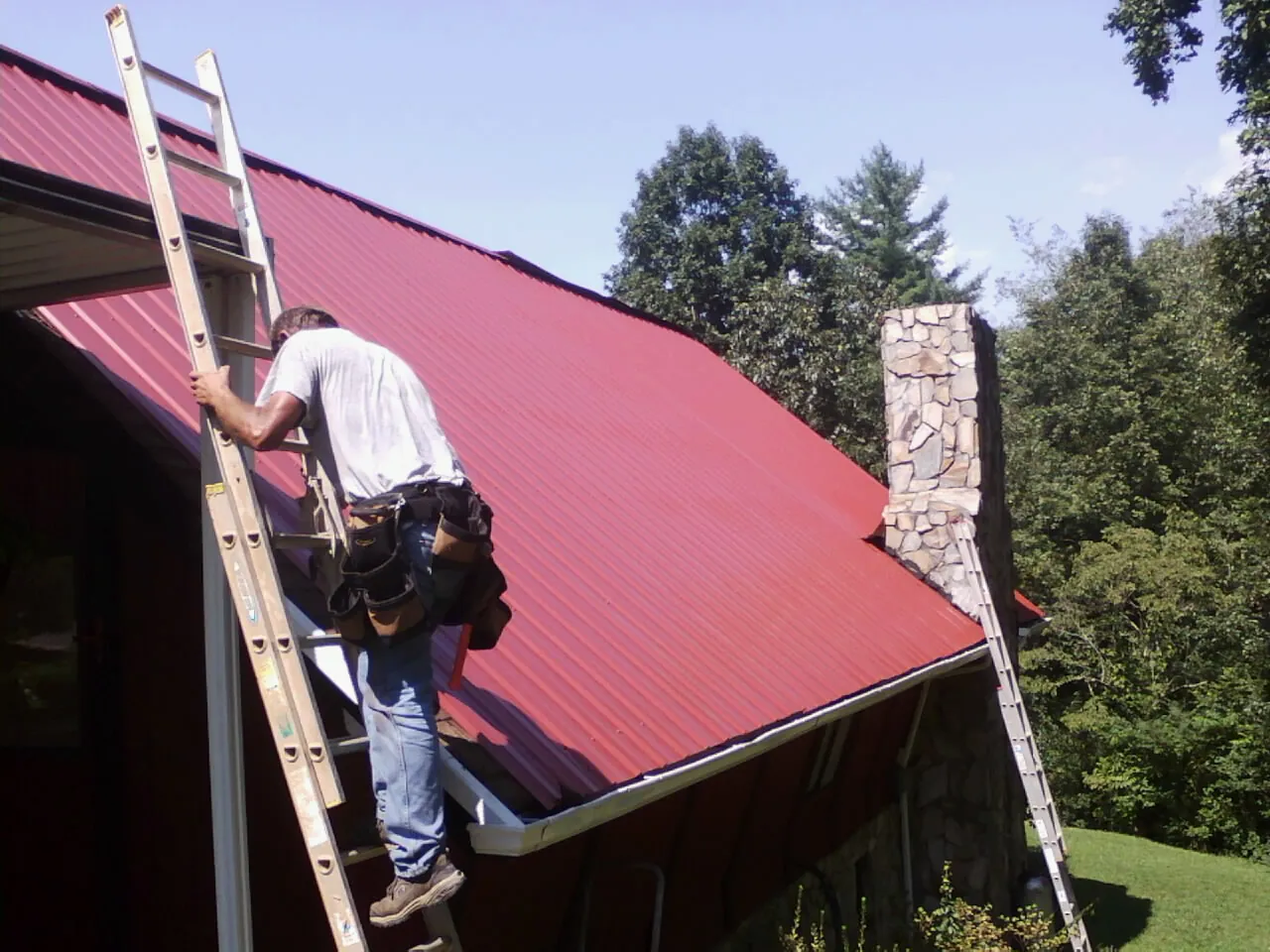Roofer on a ladder beside a red metal roof and stone chimney — Soffit & Fascia Repair by Roofing Brothers in Finger